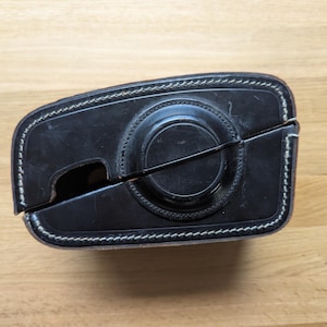 Puede incluir: Funda de cámara de cuero negro con costuras blancas. La funda tiene una abertura circular para la lente y una forma rectangular. La funda está abierta, revelando el interior. La funda está sobre una superficie de madera.
