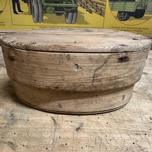 May include: A round, light brown wooden container with a flat lid and visible wood grain. The cylindrical vessel features a stepped design with a wider top section and a narrower base, showing small metal fasteners embedded in the weathered wood surface.