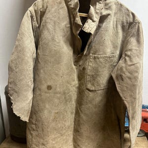 以下が含まれることがあります： スタンドカラー、フロントのハーフボタン、胸ポケットが特徴の、ベージュ色のテクスチャーのある長袖シャツ。生地には小さなシミや右袖口のほつれなど、目に見える使用感があります。
