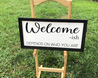 May include: A black framed sign with white text that reads "Welcome -ish Depends on who you are" on a wooden easel in a grassy area.
