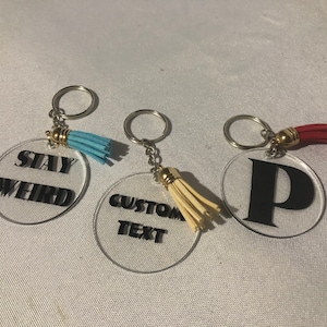 May include: Three clear acrylic keychains with different designs. The first keychain has the text "STAY WEIRD" in black lettering. The second keychain has the text "CUSTOM TEXT" in black lettering. The third keychain has the letter "P" in black lettering. Each keychain has a small tassel attached to the ring.
