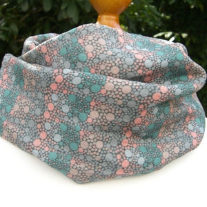 Könnte beinhalten: Ein grauer, rosa und türkisfarbener Muster-Infinity-Schal aus weichem Stoff. Der Schal ist gefaltet und auf einer Puppe ausgestellt.