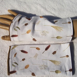 Può includere: Un paio di scaldamani in tessuto bianco con bordino marrone. Il tessuto presenta un motivo di erbe e foglie secche marroni, bianche e verdi.