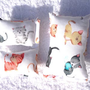 Könnte beinhalten: Vier weiße, rechteckige Kissen mit Katzenillustrationen. Die Katzen sind in verschiedenen Posen und Farben dargestellt, darunter Grau, Orange und Schwarz. Die Kissen liegen auf einer weißen, strukturierten Oberfläche.