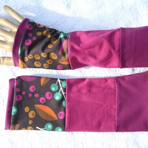 Könnte beinhalten: Ein Paar Armwärmer in Braun und Burgunderrot mit einem Blumenmuster. Die Armwärmer haben einen burgunderfarbenen Bund und einen braunen Stoff mit einem Muster aus Blättern, Beeren und kleinen Punkten.