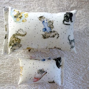 Könnte beinhalten: Zwei rechteckige Kissen mit Katzenmuster. Die weißen Kissen sind mit einem Muster aus Katzen in verschiedenen Posen und verstreuten schwarzen Punkten verziert. Die Katzen sind in Grau-, Braun- und Schwarztönen gehalten.