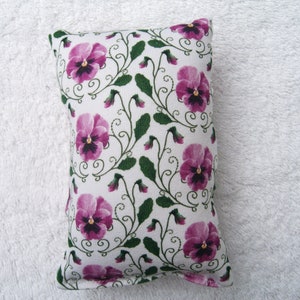 May include: A white cushion with a floral pattern of purple pansies and green leaves. The cushion is rectangular and has a soft, plush texture.