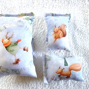 Könnte beinhalten: Drei dekorative Kissen mit winterlichen Illustrationen. Eines zeigt einen Eislaufenden Hirsch, die anderen Eichhörnchen. Die Kissen haben einen hellblauen Hintergrund mit einem winterlichen Landschaftsdesign.