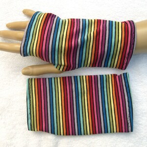 May include: Fingerless gloves with a vibrant rainbow stripe pattern. The fabric features vertical stripes in red, orange, yellow, green, blue, purple, and pink, separated by thin black lines. The gloves are shown on a mannequin hand.