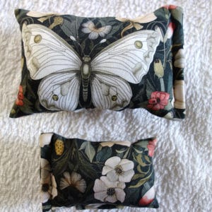 Könnte beinhalten: Zwei dekorative rechteckige Kissen mit Blumen- und Schmetterlingsmustern. Das größere Kissen zeigt einen großen weißen Schmetterling auf dunklem Hintergrund mit Blumen. Das kleinere Kissen hat weiße Blumen auf dunklem Hintergrund. Die Kissen liegen auf einer weißen strukturierten Oberfläche.