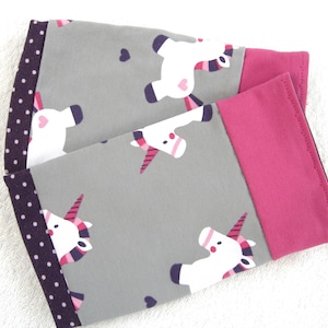 Könnte beinhalten: Ein Paar Armwärmer aus grauem und rosafarbenem Stoff mit einem Einhorn-Druck. Die Armwärmer haben einen rosa Saum und einen violetten Tupfen-Saum.