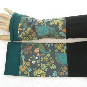 Könnte beinhalten: Armstulpen mit einem Pfau- und Blumenmuster. Der Stoff ist dunkelgrau mit türkisfarbenen, orangefarbenen und gelben Akzenten. Die Bündchen sind schwarz und grün. Diese Armstulpen sind zum Warmhalten der Arme gedacht.