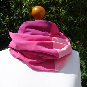 Può includere: Una sciarpa ad anello color-block caratterizzata da diverse tonalità di rosa, tra cui magenta, rosa acceso e rosa confetto chiaro. Il tessuto è drappeggiato in pieghe morbide e voluminose, creando una forma circolare su una superficie bianca.
