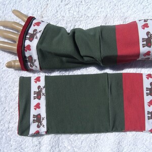 Könnte beinhalten: Ein Paar grüne Armwärmer mit rotem und weißem Besatz. Der Besatz zeigt ein sich wiederholendes Muster aus roten Herzen und braunen Rentieren.