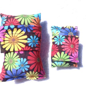 Könnte beinhalten: Zwei Sets aus Stoffartikeln mit einem farbenfrohen Blumenmuster. Das größere Set hat drei gestapelte Teile, während das kleinere Set zwei hat. Die Blumen sind in Gelb-, Rosa-, Blau- und Grüntönen auf dunklem Braun gehalten.