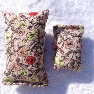 May include: Two floral patterned fabric items. The larger item is rectangular, and the smaller item is square. The fabric has a beige background with a repeating pattern of pink, red, and green flowers and dark brown stems.