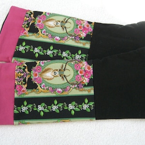 Op de afbeelding: Een paar zwarte en roze beenwarmers met een bloemen- en hertenprint. De beenwarmers hebben een zwarte stoffen manchet aan de bovenkant.