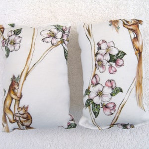 Könnte beinhalten: Zwei weiße Stoffkissenbezüge mit einem Blumen- und Eichhörnchen-Design. Die Kissen haben einen weißen Hintergrund mit braunen Ästen, rosa und weißen Blumen und braunen Eichhörnchen.