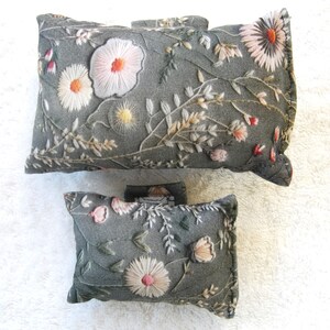 May include: Two grey floral embroidered pillow covers with a delicate floral pattern. The larger pillow cover has a white background with pink and orange flowers, while the smaller pillow cover has a darker grey background with white and pink flowers.
