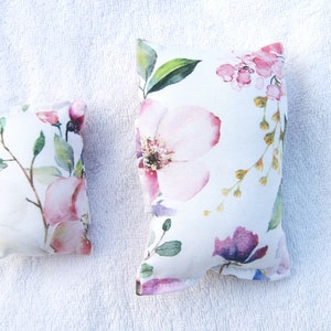 Könnte beinhalten: Zwei Dekokissen mit Blumendruck, eines größer als das andere, mit weißem Hintergrund und rosa, violetten und grünen Aquarellblumen.