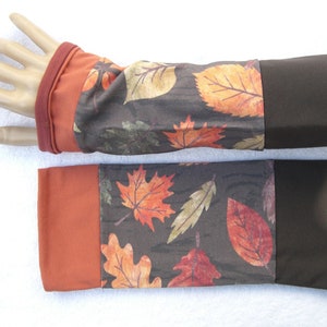 May include: A pair of brown and orange arm warmers with a leaf print. The arm warmers have a brown cuff at the top and a rust colored cuff at the bottom.
