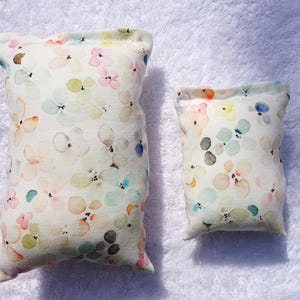 Könnte beinhalten: Zwei rechteckige Kissen mit floralem Muster im Aquarellstil. Die Kissen haben einen cremefarbenen Hintergrund mit bunten Blumenmustern in Rosa, Blau, Gelb und Grün. Das größere Kissen ist etwa 20 cm lang, und das kleinere etwa 13 cm lang.