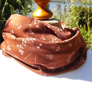 Puede incluir: Una bufanda de seda marrón con un estampado floral blanco. La bufanda está doblada y drapeada sobre un maniquí.