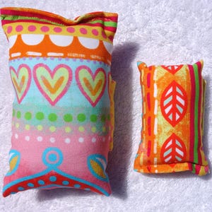 Könnte beinhalten: Zwei bunte Stoffkissen. Eines ist rechteckig mit einem Muster aus Herzen, Punkten und Wirbeln in Rosa, Blau, Orange und Gelb. Das andere ist kleiner, mit einem Blattmuster in Orange, Gelb und Rot.