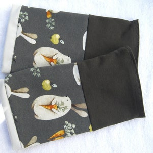 May include: A pair of gray and brown oven mitts with a white rabbit and carrot pattern. The mitts have a white trim and a dark brown back.