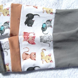 Könnte beinhalten: Ein Paar graue, braune und weiße Beinwärmer aus Stoff mit einem sich wiederholenden Muster aus Comic-Katzen. Die Katzen sind in verschiedenen Posen und Farben, darunter grau, schwarz, rosa und gelb.