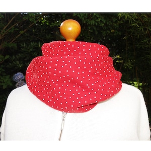 Può includere: Sciarpa rossa a pois bianchi intorno al collo. La sciarpa è realizzata in tessuto morbido ed è abbinata a un maglione bianco. Un bottone di legno è visibile nella parte superiore della sciarpa.