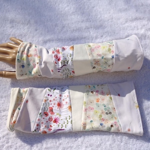 May include: A pair of white and floral patterned arm warmers. The arm warmers are made of fabric with sections of white and sections with a floral pattern in pink, yellow, and blue. The arm warmers are displayed on a mannequin hand.