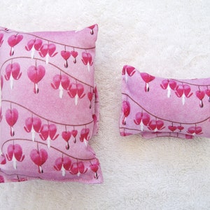 Könnte beinhalten: Zwei rosa Stoffkissen mit einem Blumenmuster aus Herzblumen. Das größere Kissen ist rechteckig und das kleinere Kissen ist ein langes Rechteck.