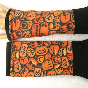 Könnte beinhalten: Ein Paar Armwärmer in Schwarz und Orange mit einem Kürbisgesichtsmuster. Die Armwärmer haben schwarze Bündchen.