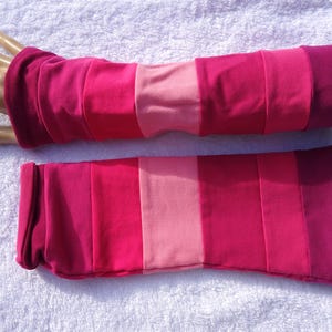 Könnte beinhalten: Ein Paar Armstulpen in Rosa- und Magentatönen. Die Armstulpen aus Stoff haben ein gestreiftes Design mit verschiedenen Rosatönen. Ein Armstulpen wird auf einer Mannequinhand gezeigt.