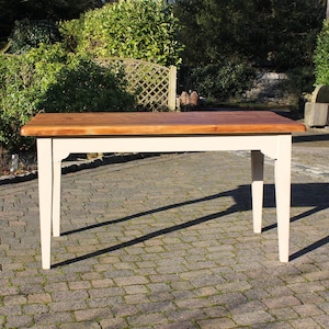 May include: A rectangular wooden table with a light brown top and cream-coloured legs. The table is approximately 122 cm long and 61 cm wide, with a classic design. The table is outdoors on a brick surface.
