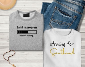 May include: Two gray and white sweatshirts with religious-themed text. The gray sweatshirt has the text "Saint in progress" with a progress bar and the text "holiness loading..." The white sweatshirt has the text "striving for Sainthood" in gold lettering.