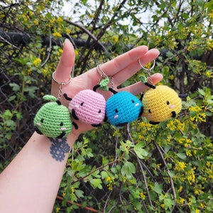 Puede incluir: Cuatro abalorios de llavero de ganchillo en forma de una pequeña fruta verde, rosa, azul y amarilla con una pequeña hoja verde en la parte superior. Cada abalorio tiene una cara bordada en negro con una simple sonrisa.