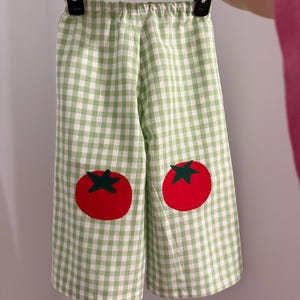 May include: Green and white gingham patterned pants with two large red tomato appliqués. The pants are hanging on a black hanger. The tomatoes have green stems and are centered on the knees.