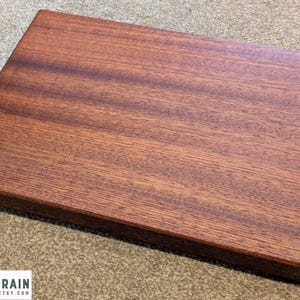May include: A rectangular wooden cutting board with a rich, reddish-brown hue. The wood grain is visible, adding texture to the surface. The board has a smooth, finished appearance, suggesting it's ready for use in the kitchen.