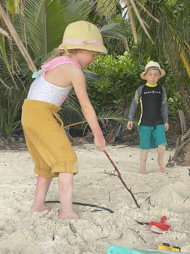 May include: Two children are playing on a sandy beach. A girl wearing a yellow and white swimsuit, a straw hat with a pink ribbon, and yellow trousers holds a stick. A boy wearing a black rash guard with the text "Aqua Lung" and green swimming trunks stands nearby.