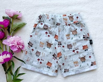 May include: Pale blue baby shorts with a charming woodland animal design. The shorts display a repeating pattern of foxes, deer, bears, and rabbits, with small red mushrooms and floral details. They have an elasticated waistband and pockets.