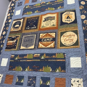 May include: A blue and brown patchwork quilt with a camping theme. The quilt features various designs, including a coffee cup, a camper van, a lake, and a mountain scene. The quilt is made of different fabrics and has a quilted texture.