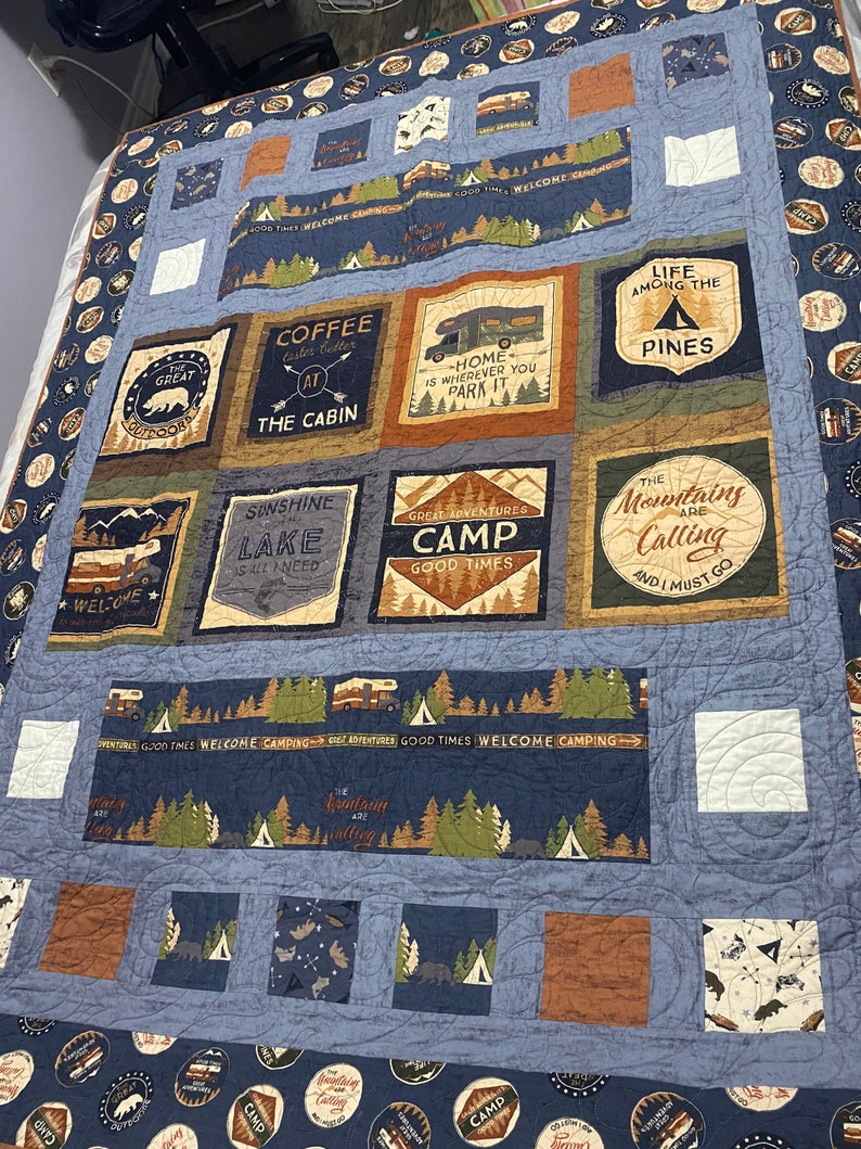 May include: A blue and brown patchwork quilt with a camping theme. The quilt features various designs, including a coffee cup, a camper van, a lake, and a mountain scene. The quilt is made of different fabrics and has a quilted texture.
