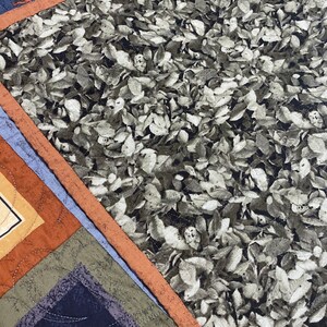 May include: A close-up of a quilt with a brown, orange, and blue patchwork design. The quilt features a pattern of leaves in shades of grey and green.