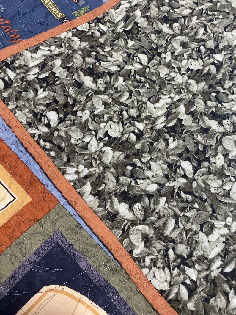 May include: A close-up of a quilt with a brown, orange, and blue patchwork design. The quilt features a pattern of leaves in shades of grey and green.