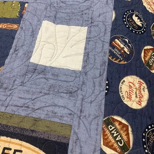 May include: A blue and white patchwork quilt featuring various patterns and colours, including a white patch with a leaf design, a blue patch with a swirl design, and a brown patch with a star design. The quilt also features several circular patches with text and images related to camping, such as "Good Times, Great Adventures, Camp" and "The Mountains Are Calling".