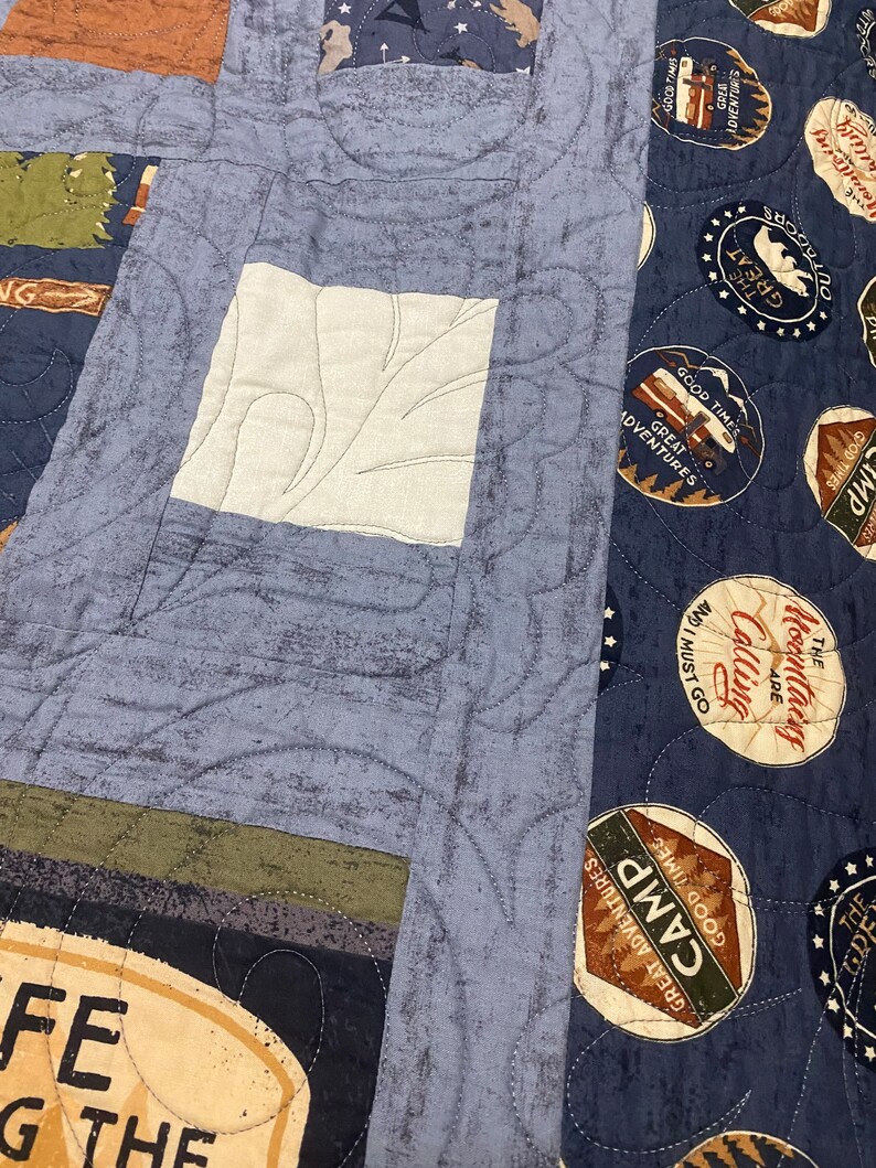 May include: A blue and white patchwork quilt featuring various patterns and colours, including a white patch with a leaf design, a blue patch with a swirl design, and a brown patch with a star design. The quilt also features several circular patches with text and images related to camping, such as "Good Times, Great Adventures, Camp" and "The Mountains Are Calling".