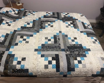 May include: A white quilt with a geometric pattern in shades of blue, grey, and black. The quilt is made up of squares and rectangles, and the pattern is repeated throughout the quilt.