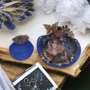 Puede incluir: Un adorno azul en forma de concha y un colgante azul en forma de calabaza con una ilustración de bruja. Ambos están adornados con detalles dorados y rojos. El colgante presenta un diseño de mariposa y flores.